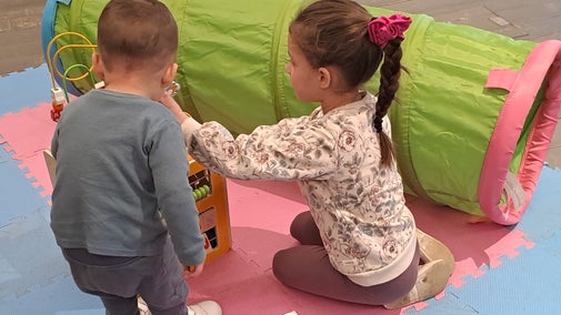 Two young children playing with a wooden toy on pink and blue mats with a colourful play tunnel next to them.